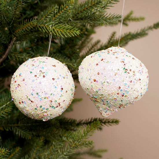 Two glittery white Christmas ornaments hanging on a tree branch.
