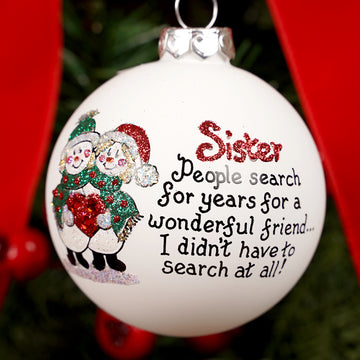 Christmas ornament with text and sister snowman illustration, hanging against a red background.