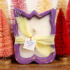 Decorative candle with a purple wavy holder in front of colorful bottlebrush trees.
