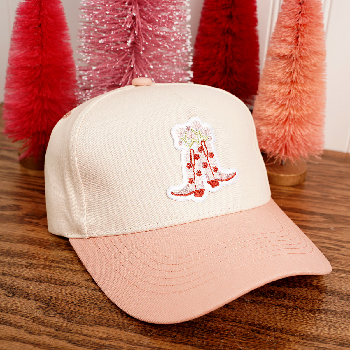 Beige and pink cap with a red boot design on a wooden surface with Christmas trees in the background