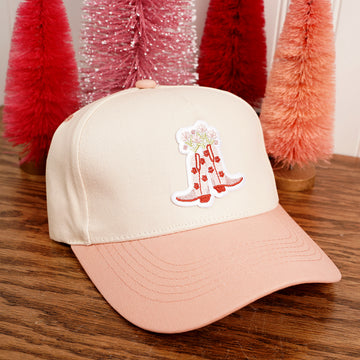 Beige and pink cap with a red boot design on a wooden surface with Christmas trees in the background
