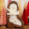 Candle shaped like a boot with a wooden holder, surrounded by festive bottlebrush trees.